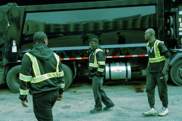 Three people wearing high-visibility jackets stand near a large truck. They appear to be working or taking a break. Urban buildings are reflected on the side of the vehicle.