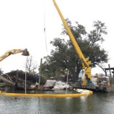 A large industrial crane and excavator work to remove debris from a body of water, with workers supervising the operation. A yellow barrier surrounds the area.