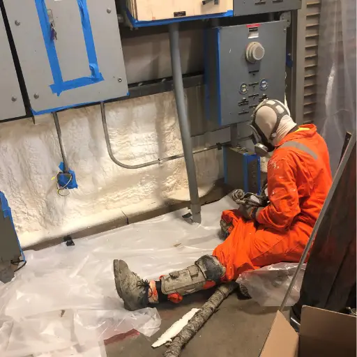A person in an orange suit and hard hat working on equipment in an industrial setting, seated on a plastic-covered floor.