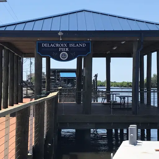 An empty pier with a blue metal roof labeled "Delacroix Island Pier" extends over water under a clear blue sky.