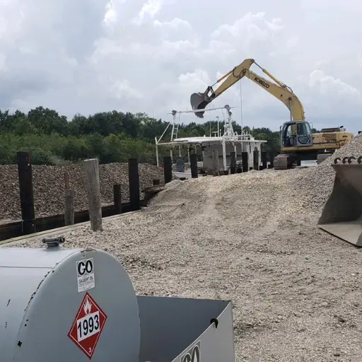 Excavator loading gravel onto a large boat docked at a gravel-covered port with a fuel tank labeled "1993" in the foreground.