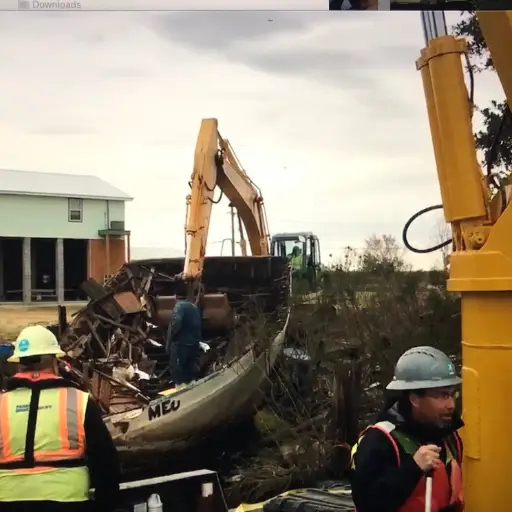 Construction workers use heavy machinery to clear debris from a grounded boat near a house.