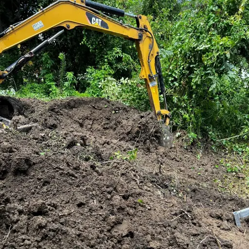 A yellow excavator with a claw attachment is digging into a dirt mound surrounded by green foliage.