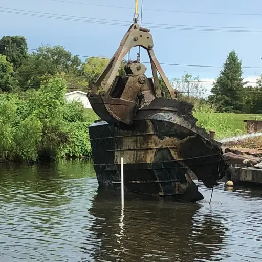 An excavator claw lifts a large chunk of debris from a body of water, with greenery and a structure visible in the background.