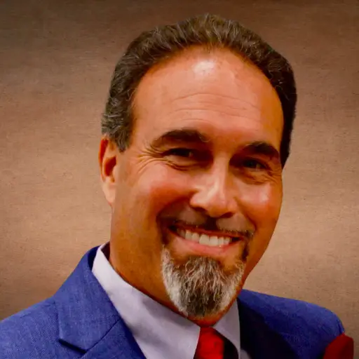 A man in a blue suit and red tie smiles against a brown background.
