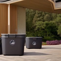 Three black plastic trash bins with recycling symbols on a paved outdoor area, under an overhang. Background features trees and shrubbery.